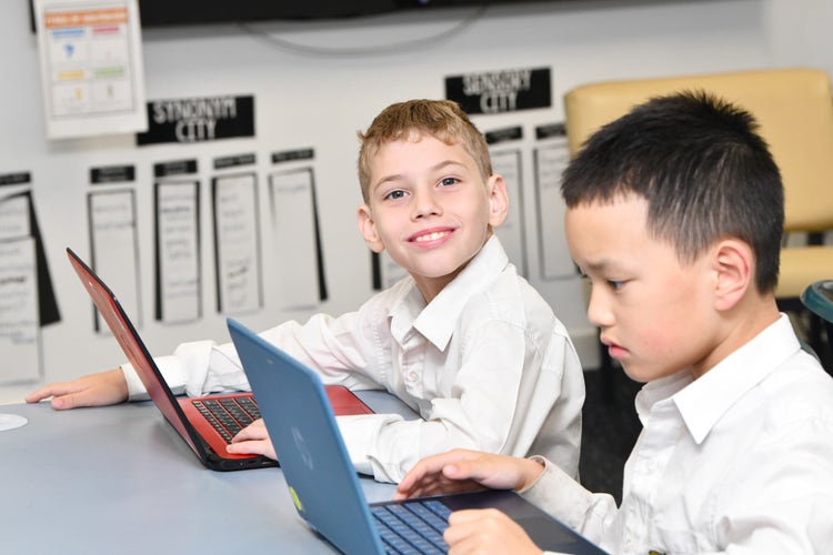 Two students sitting at a desk working on a laptop. One student is looking at the camera and the other at the screen.