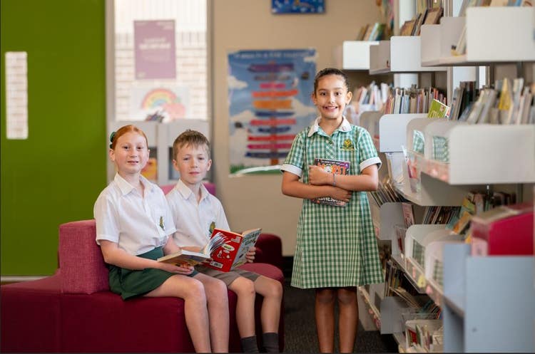 Three students in a library holding books. Two sitting and one standing. All looking at the camera.