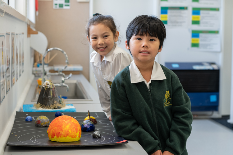 Boy standing in the foreground facing the camera with a girl looking over his shoulder. Both leaning against a bench and a science project of the solar system sitting on the bench.