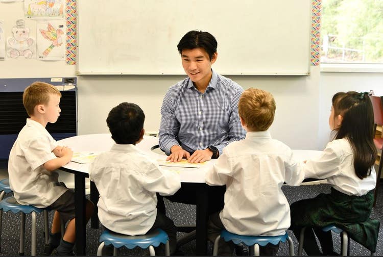Teacher sitting at a jellybean shaped table with four students sitting on the other side looking at him.