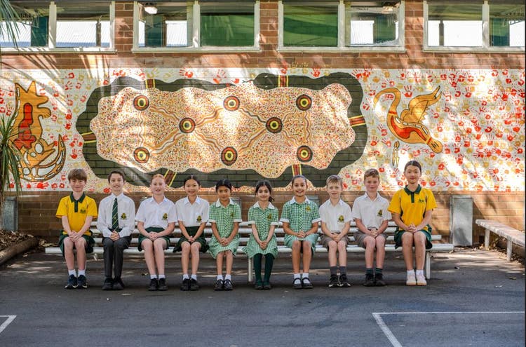10 students sitting on a silver seat in front of an Aboriginal Art mural.