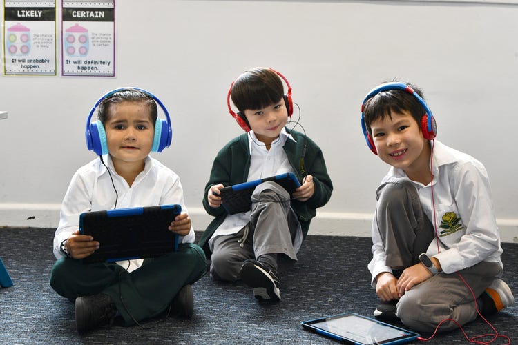 3 Year 1 students sitting on the floor holding iPads with headphones on.