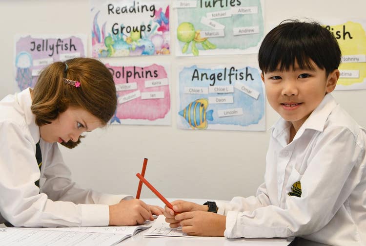 Girl on the left looking down writing with boy seated opposite her holding a pencil and looking at the camera.