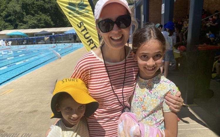 Mother standing between 2 daughters at the side of a swimming pool.