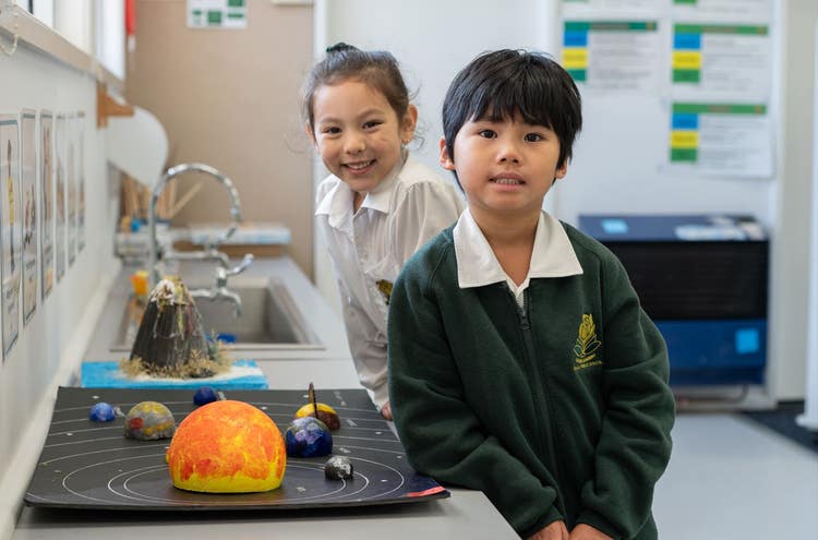 Two students leaning against a bench both facing the camera. Science project sitting on the bench.